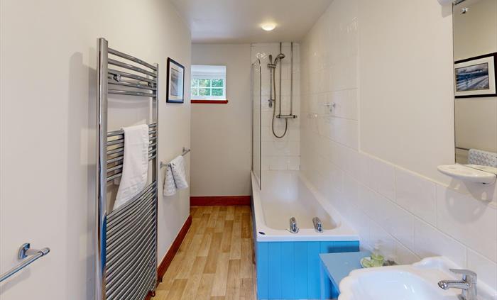 A bright and practical family bathroom featuring a full-sized bath with overhead shower, wooden flooring, a large wall mirror, and chrome towel rails.